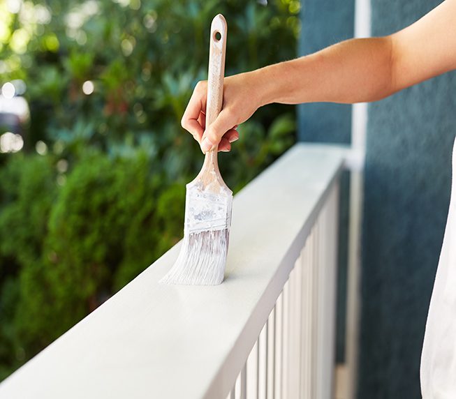 Human hand painting a house deck handrail, doing home improvement by refreshing the paint of the wooden structure at the house entry. Fresh wet white paint.